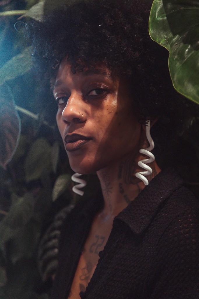 Photograph in color of a woman with brown skin, she has dark curly hair, and she is wearing white sculptural earrings. She is facing the camera and is photographed against a backdrop of green plant leaves.