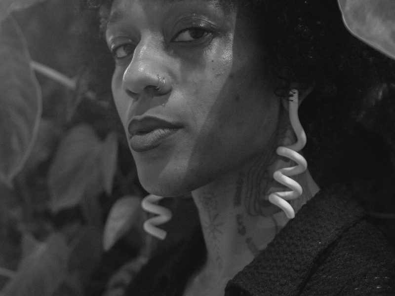 A woman stares into the camera. Her face is framed by her hair and she wears large, spiral shaped earrings
