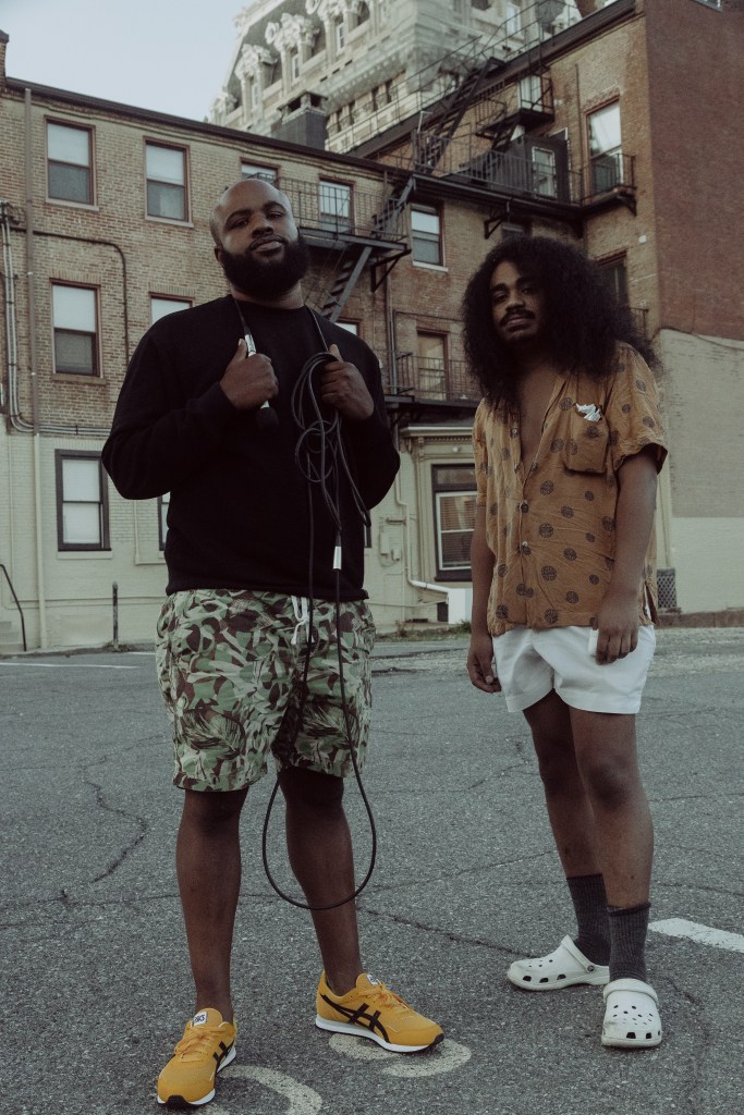 A color photograph of two men with brown skin. One is wearing a black long sleeved shirt, camo shorts, and yellow sneakers. He has a bald head and is holding a microphone. The other figure is wearing a polka dot shirt, white shorts, and white crocs, and he has long curly hair. They are standing in front of brick buildings.