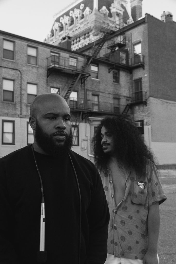 Black and white photograph of two men with brown skin. One is wearing a black long-sleeved shirt and has a bald head, the other figure is wearing a polka dot shirt and he has long curly hair. They are standing in front of brick buildings.