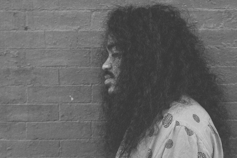 A man with brown skin is looking away from the camera, facing the left side of the image, he is photographed in front of a brick wall, he has curly black hair, a mustache, and is wearing a patterned shirt. He is pictured with his eyes closed.