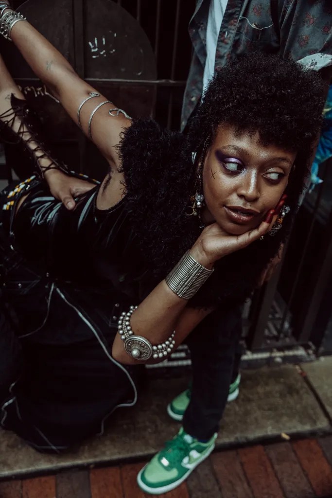 Safra is pictured looking away from the camera, wearing silver jewelry on both arms and a black garment. Safra has curly hair and brown skin.
