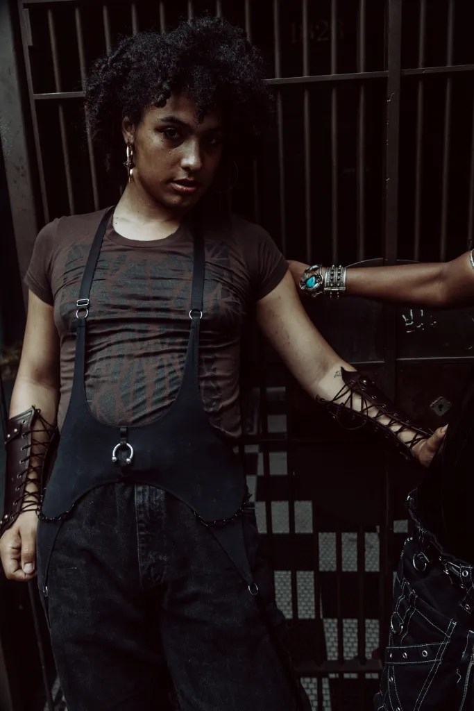Proxy is wearing an olive colored top and black pants with suspenders. Proxy is looking at the camera and is also waearing a leather sleeve and earrings, Proxy has curly hair and earrings.