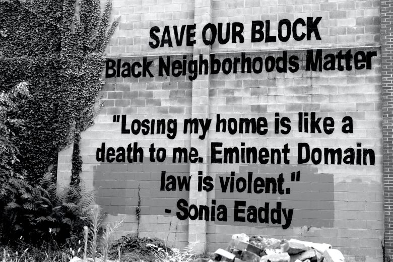 A black and white photo of a brick wall—the side of a home owned by the Eaddys in Poppleton. Painted in large photos on the wall "SAVE OUR BLOCK. Black Neighborhoods Matter. "Losing my home is like a death to me. Eminent Domain law is violent."-Sonia Eaddy
In front of the wall and mural is debris and overgrown plants.