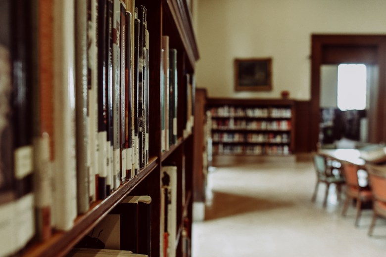 A library is displayed. There are wooden bookshelves full of books and in the background out of focus is a large table and chairs. 