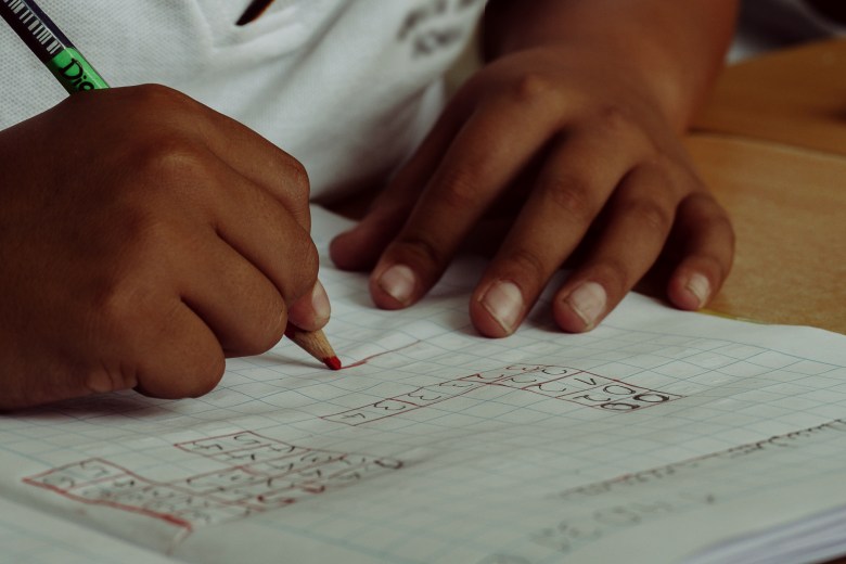 A child's hands are displayed as they work on a math problem. They are holding a red colored pencil and writing in notebook. 