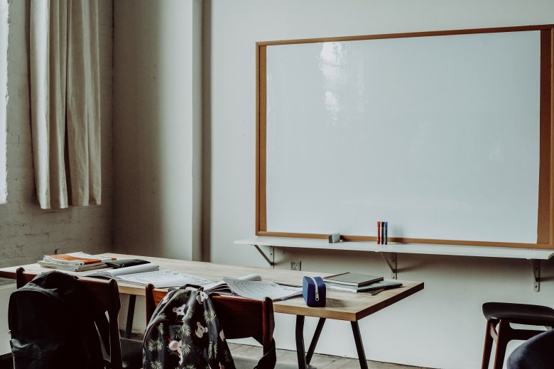 A classroom is displayed, there is a wooden desk with supplies and books, two backpacks hang on chairs, and there is a a large blank white board. 