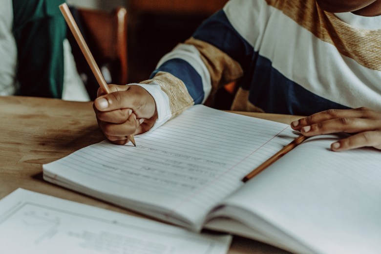 A photograph of a child holding a pencil and writing in a lined notebook at a wooden desk. The childs face is out of the frame and they are wearing a striped long sleeve shirt. 