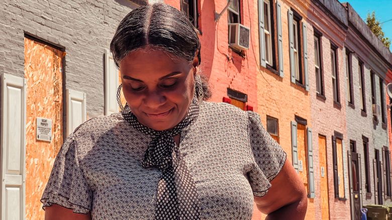 Photo of Sonia Eaddy. She is a Black woman wearing a gray and black shirt with a black and white tie. She stands behind boarded up homes of varying colors.