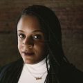 An image of a Black woman with braids. She is wearing a white shirt underneath a black jacket.