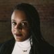 An image of a Black woman with braids. She is wearing a white shirt underneath a black jacket.