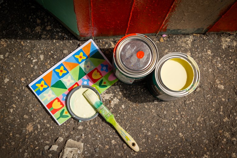 A paper with a mural plan on the ground with paints cans nearby and a used paint brush.