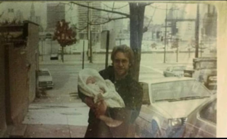 A vintage photograph of a man holding a baby. He is wearing a coat, the baby is wrapped in a white blanket. 