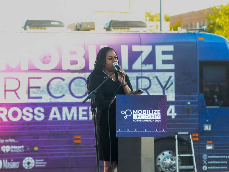 A woman stands at a podium outside. She standing in front of a bus with the words "mobilize recovery across America" printed on it.