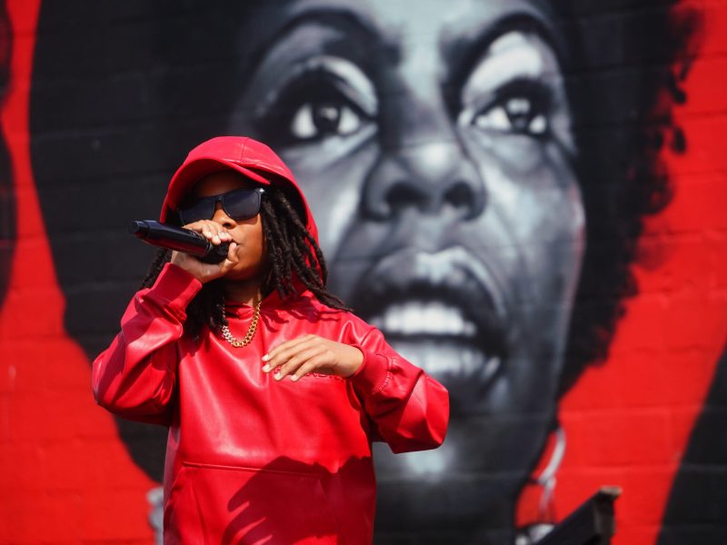 A person with brown skin raps into a microphone. They are dressed all in red and have dark locs. They stand in front of a red mural with a woman's face painted on it.