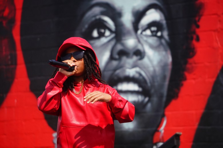 A person with brown skin raps into a microphone. They are dressed all in red and have dark locs. They stand in front of a red mural with a woman's face painted on it.
