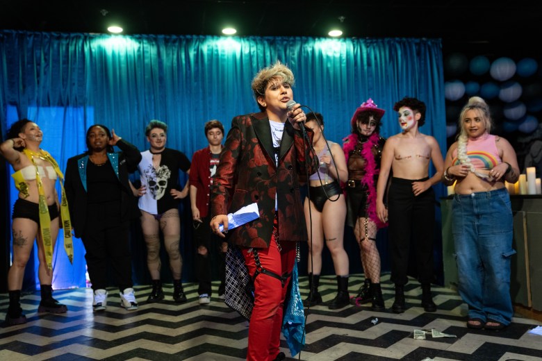 A host, wearing a red blazer and red pants, stands in front of a line of performers at a drag king and dragster showcase.