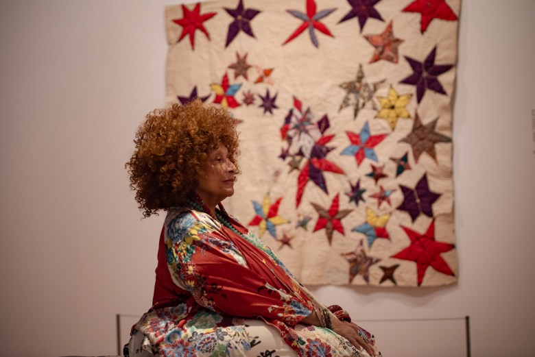 photo of a woman with brown skin sitting in front of a quilt
