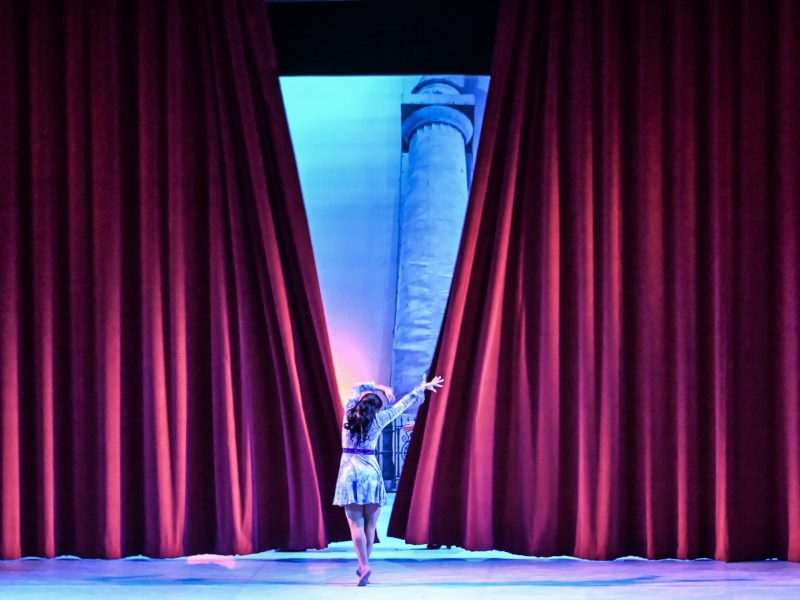 A dancer with her back to the camera. Red stage curtains open in the distance.