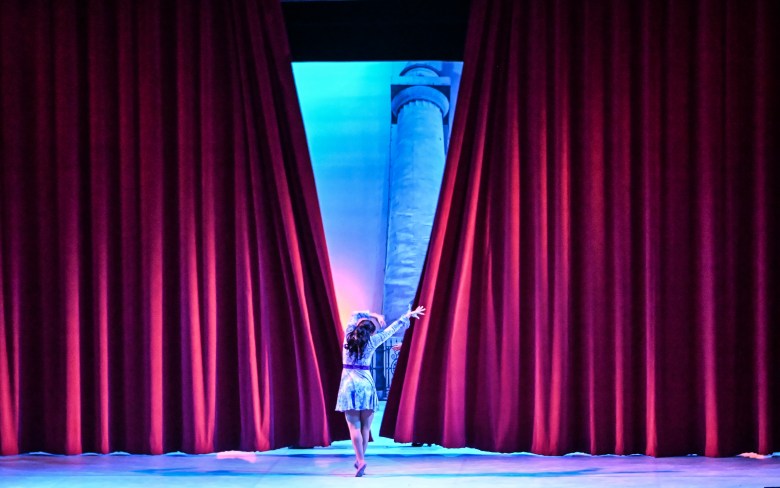 A dancer with her back to the camera. Red stage curtains open in the distance.