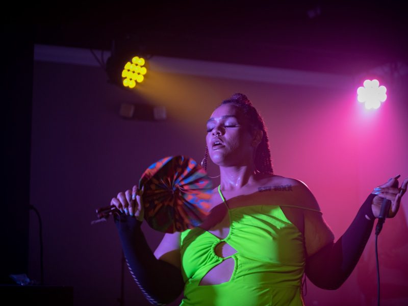 A person wearing a green top stands fanning themselves, they're in a nightclub and lights are shining on them.