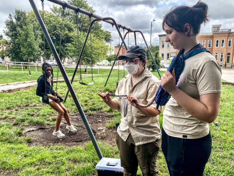 Two people wearing tan shirts and dark pants examine research equiptment. A young person on a swing looks on in the background.
