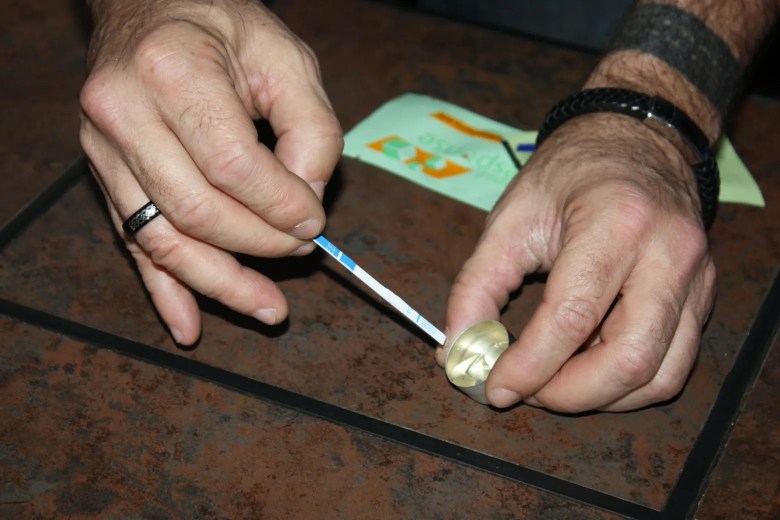 A photo of a man holding a fentanyl testing strip and dipping it into his drugs in a cooker. He has a ring on one finger on his left hand and a bracelet and tattoo on the other.