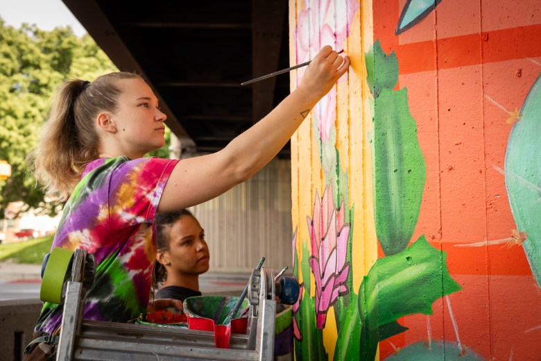 Two muralists, Sammi Seezox and Chelsea Henery, are painting plants on one of the pillars under the Jones Falls Expressway ahead of Artscape.
