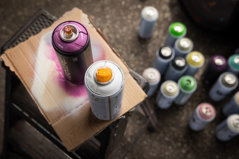 Two spray paint cans in focus on top of a piece of cardboard on a step ladder, with fifteen spray paint cans on the ground below out of focus.