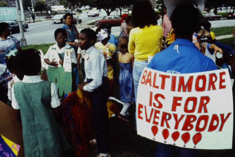 Color photograph of a group of people at an arts festival. A sign says "Baltimore Is For Everybody!!!"
