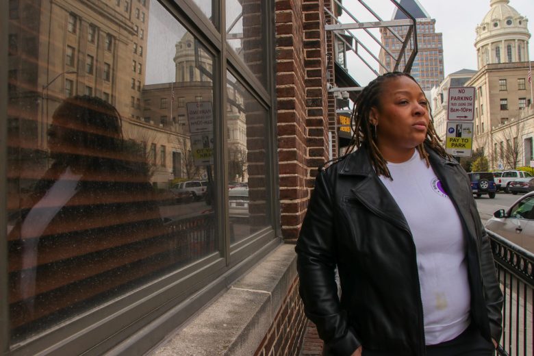 A person with brown skin wearing a black leather jacket and purple top leans against a building