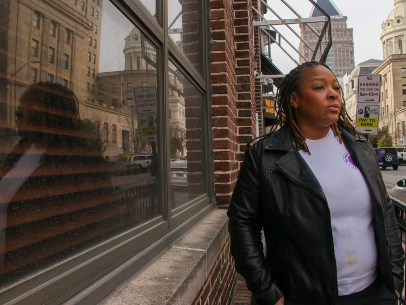 A person with brown skin wearing a black leather jacket and purple top leans against a building