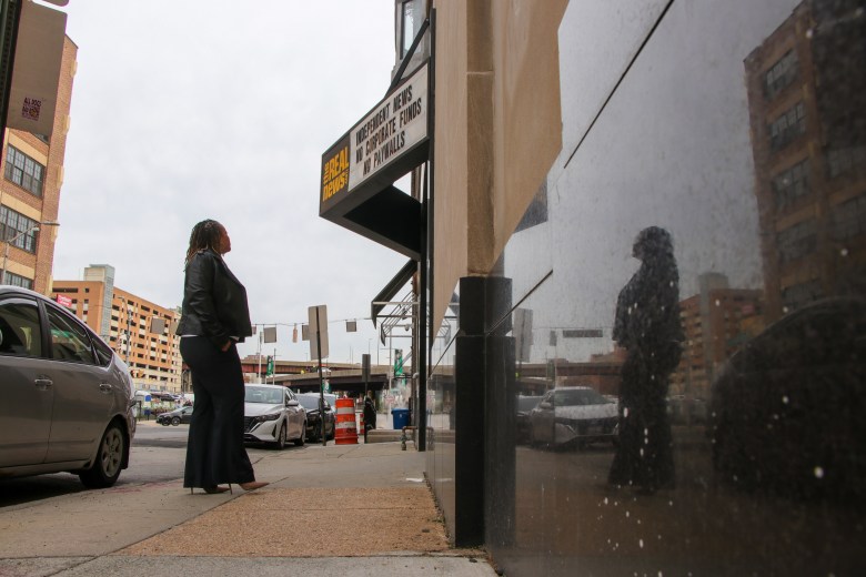 A woman stands outside under a sign for The Real News Network. She is looking away from the camera.