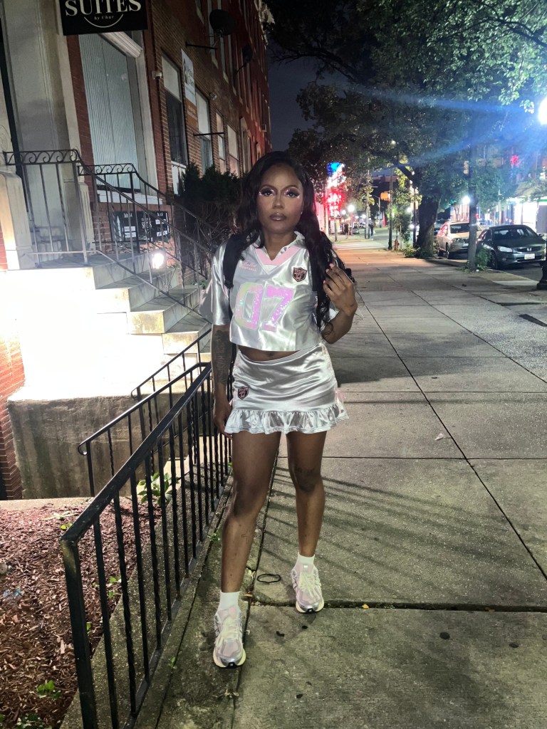 A photo of Yaira Wang standing on a street sidewalk. Wang wears a cropped silver jersey with a matching silver skirt. 