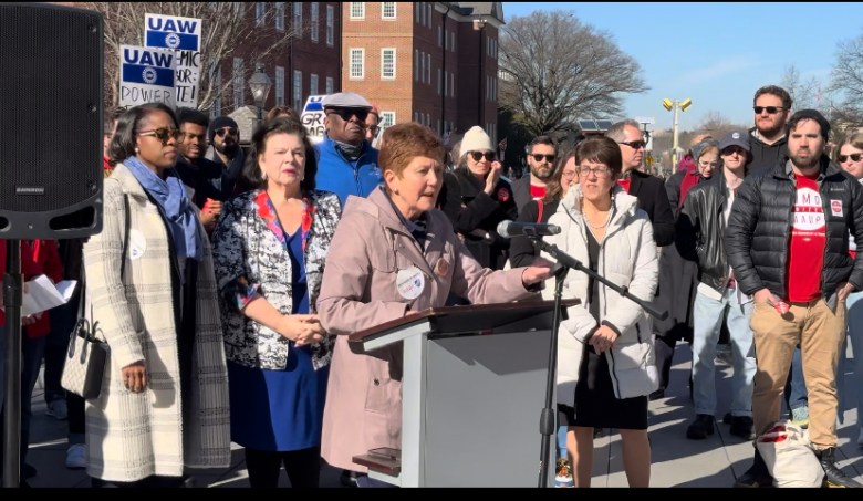 A woman with red hair and a tan coat speaks at a podium. She is surrounded by other people.