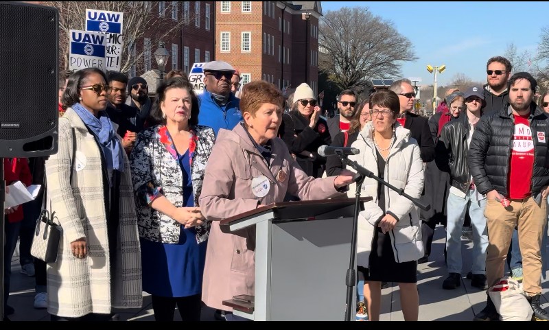 A woman with red hair and a tan coat speaks at a podium. She is surrounded by other people.
