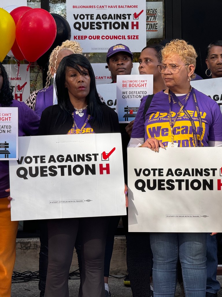 Two women hold white signs that read "vote against question H"