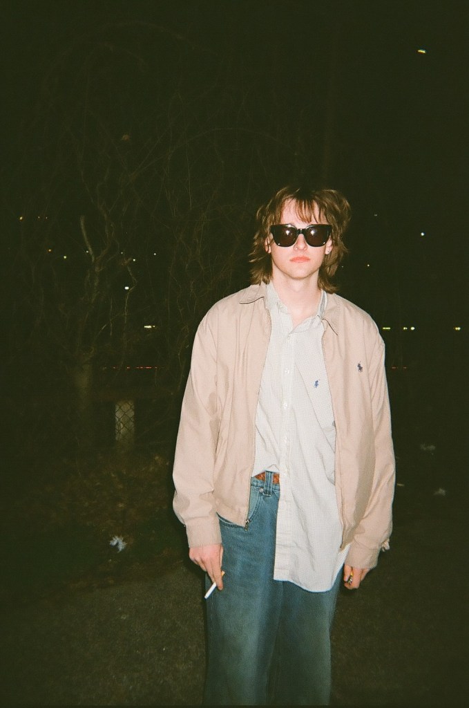 A person stands looking at the camera. They have short brown hair and are wearing sunglasses. They have on a light brown jacket over a light-colored button-down shirt and jeans. They are holding a cigarette.