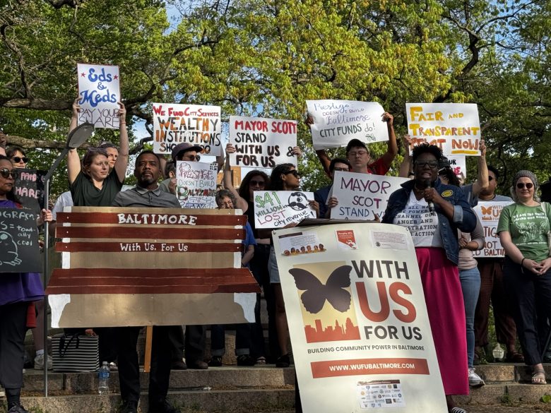 A woman wearing a denim jacket, white top, and pink pants speaks in front of a ccrowd holding signs.