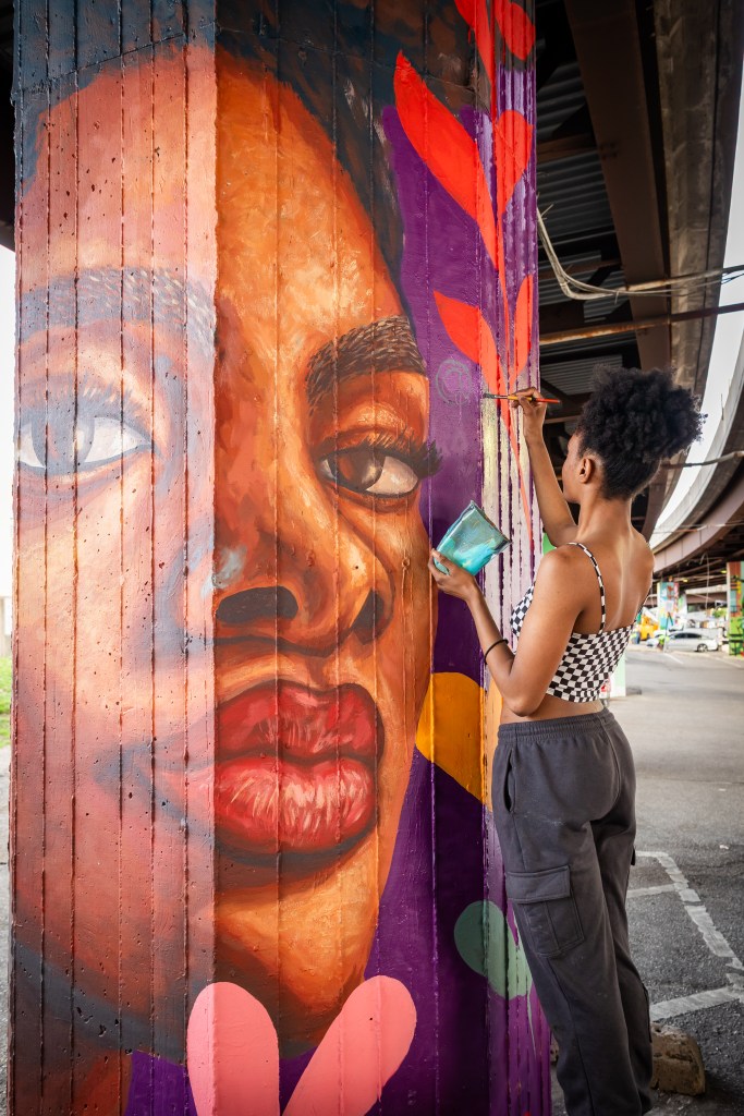 Artist Jada McAliley paints a large portrait of a Black woman on a pillar under the Jones Falls Expressway.
