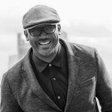 A black and white photo of a Black man wearing a cap, glasses, a button down shirt, and a blazer.