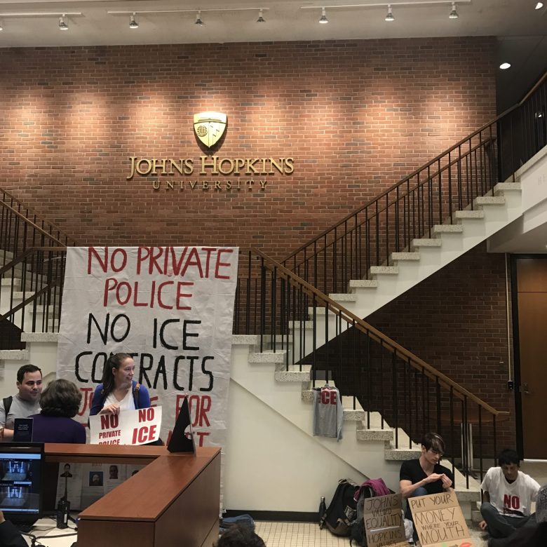 Protesters at Johns Hopkins University. People sit in a room with signs. A large white sign reads: "no private police no ice contracts."