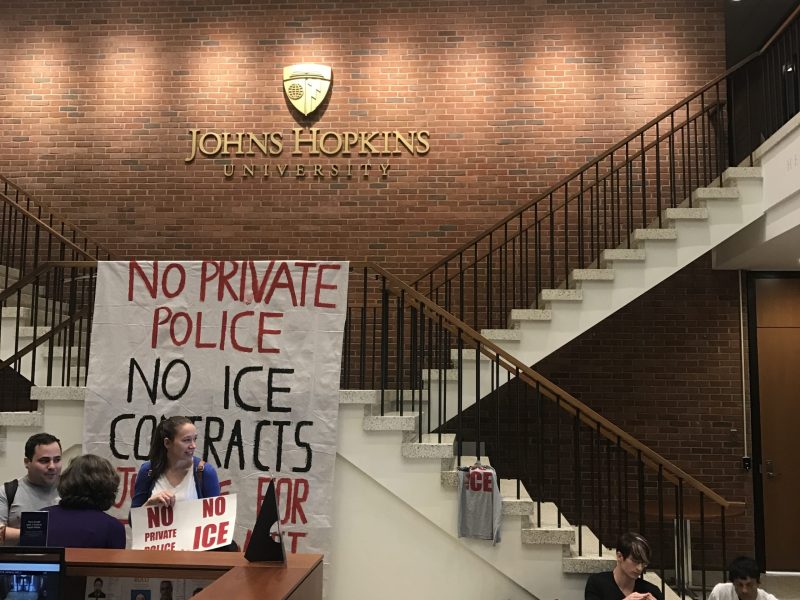 Protesters at Johns Hopkins University. People sit in a room with signs. A large white sign reads: "no private police no ice contracts."