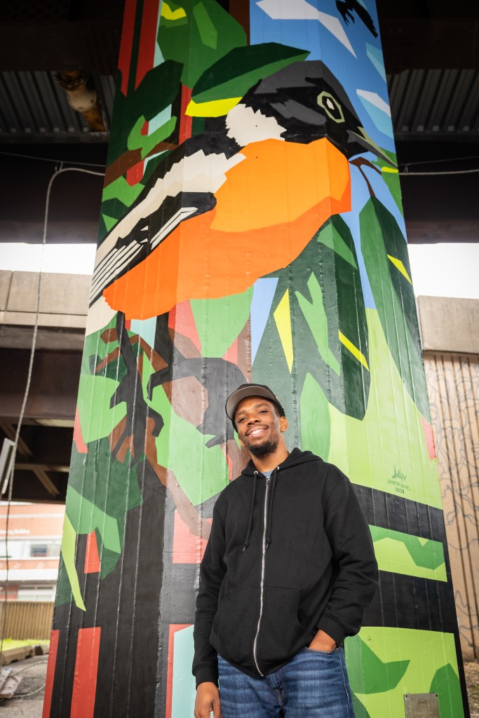 Artist Jordan Lawson stands in front of his mural featuring plants and a geometric representation of an Oriole bird.