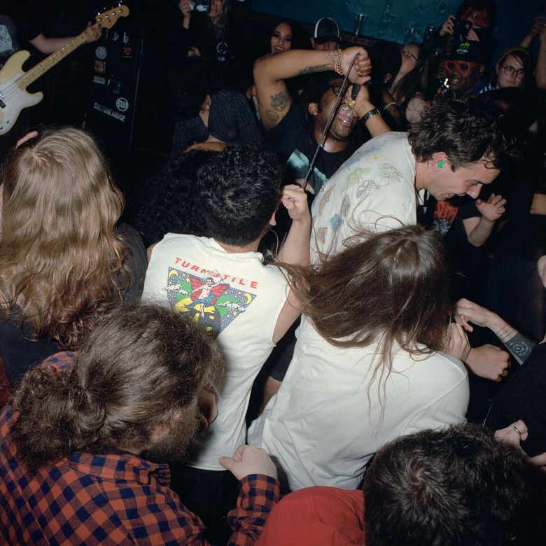 A photograph of a mosh pit with the lead singer of Soul Glo singing. He has mouth open, screaming a lyric, the crowd is excited and smiling, one audience member is wearing a cut off t-shirt that says Turnstile.