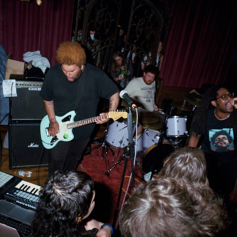 Soul Glo performing at Holy Frijoles in front of a crowd. The lead singer is wearing a T-shirt that has the singer The Weeknd on it, the guitar player is playing a blue guitar. The faces of the crowd are obscured as they look at the band.