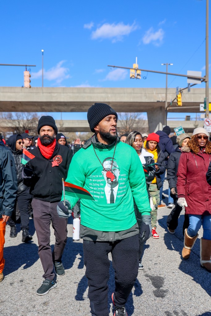 A man wearing a bright green shirt over a coat holds a Pan-African flag and walks in a parade.