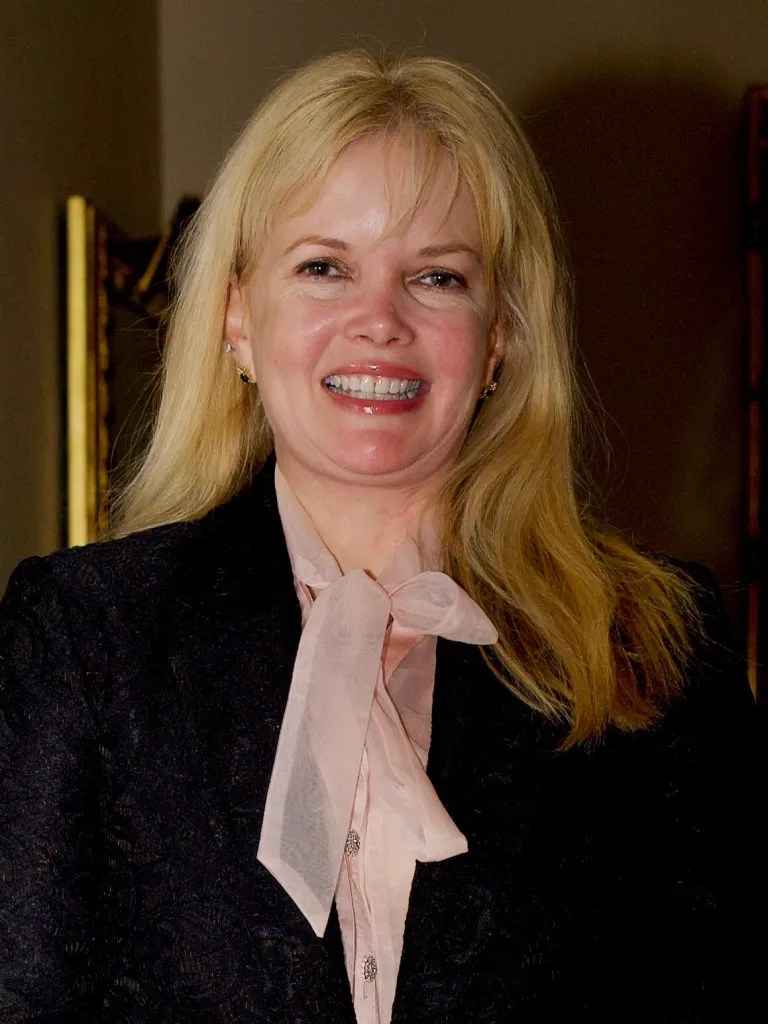 A photo of a smiling woman. She wears a pink blouse and a black blazer.