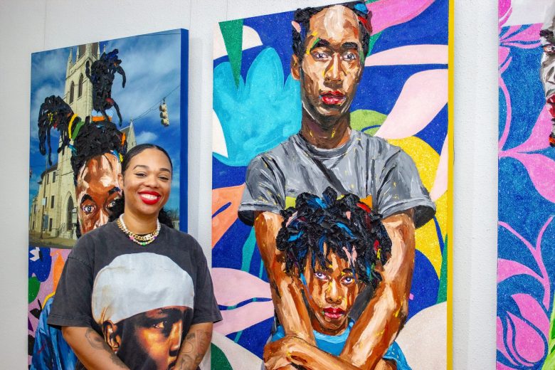 photo of an artist with brown skin, she poses in front of her paintings