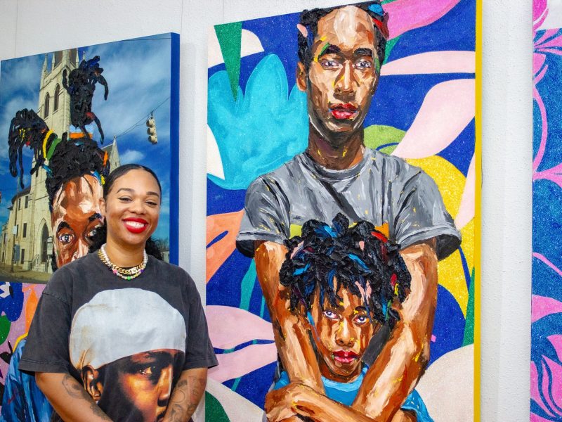photo of an artist with brown skin, she poses in front of her paintings
